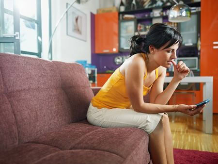 Woman in a yellow top sits on a sofa, looking at her smartphone with a thoughtful expression. Brightly colored kitchen cabinets are visible in the background.