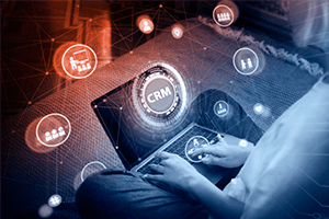 Person using a laptop with CRM software interface, surrounded by digital icons representing business and technology concepts on a network-like background.