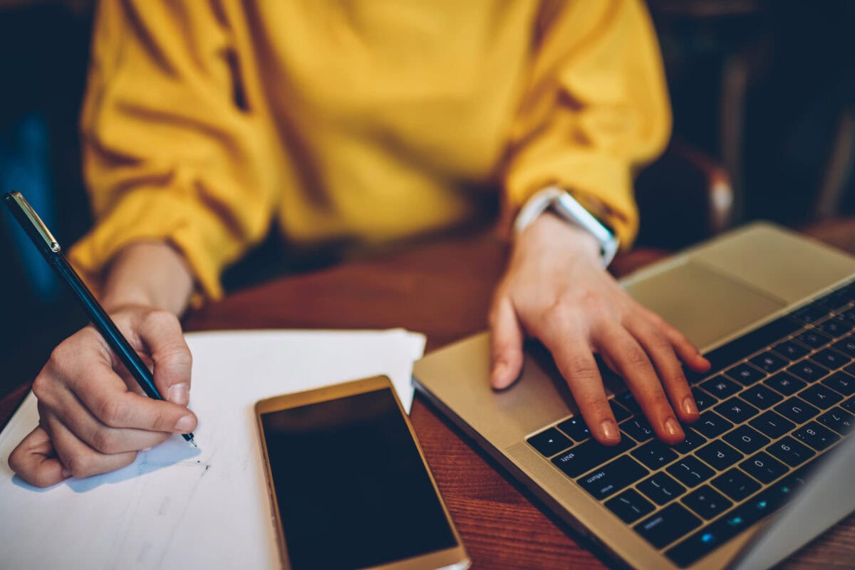 Cropped image of female hands with pen writing notes for course work using laptop computer, young female student studying at table with smartphone device for your internet content or website