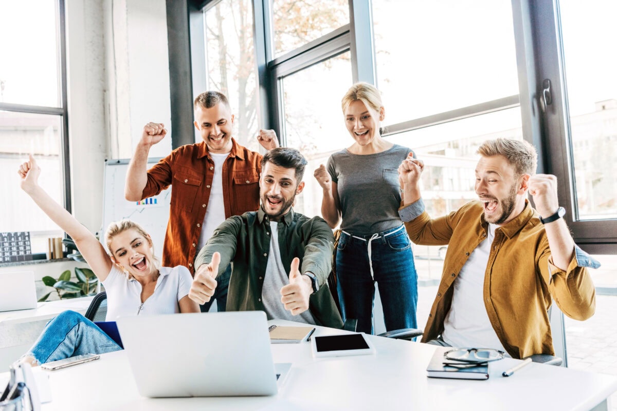 Group of young successful entrepreneurs celebrating victory and showing thumbs up at office and looking at camera