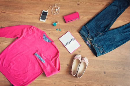 Pink sweater, blue jeans, pink wallet, pink floral shoes, smartphone with earphones, earrings, and a notebook on a wooden floor.