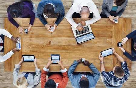 People seated around a wooden table using various electronic devices, including laptops, tablets, and smartphones.